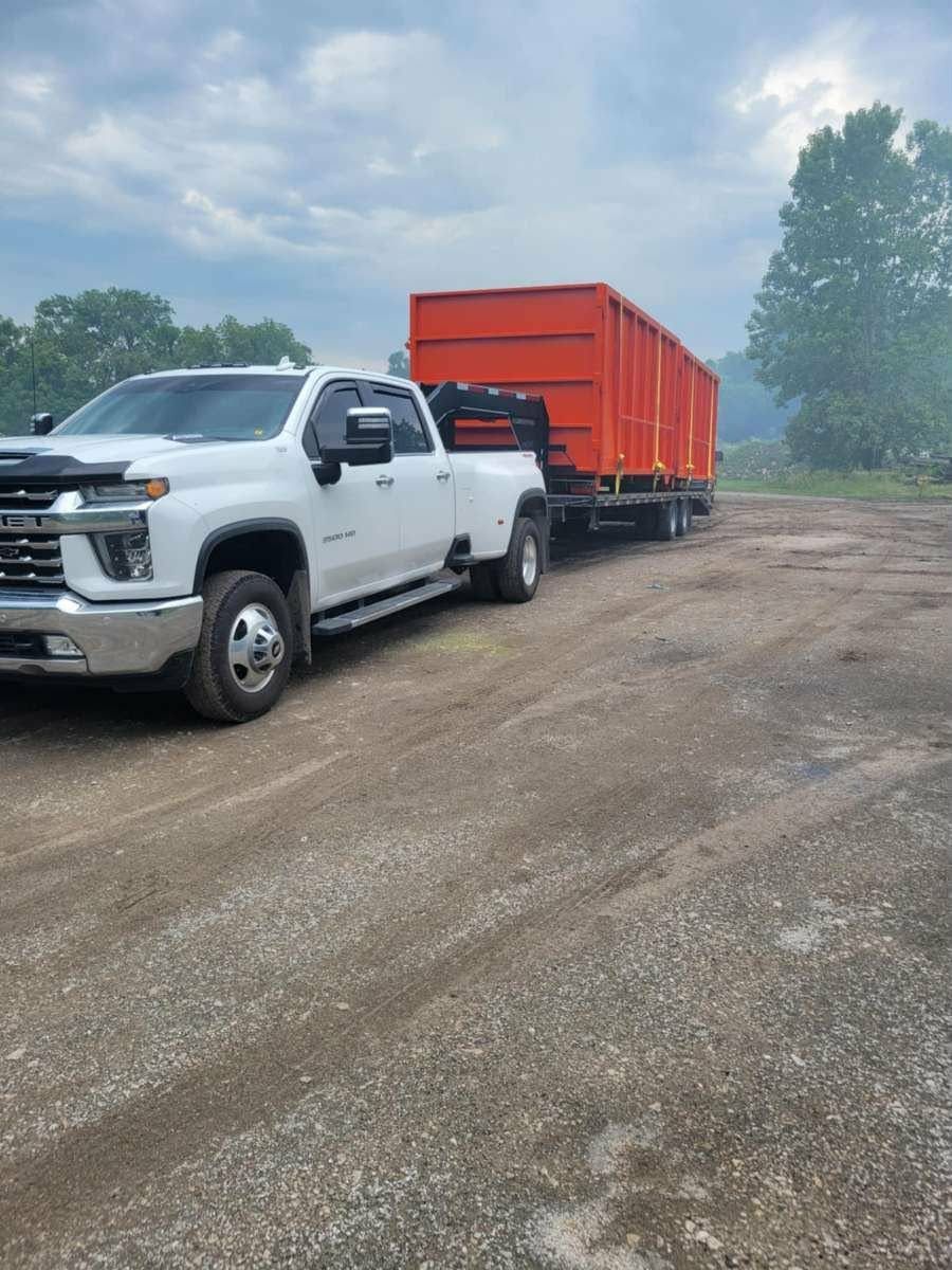 A white dually pickup truck towing a flatbed trailer with a large orange dumpster on a dirt lot under a cloudy sky.