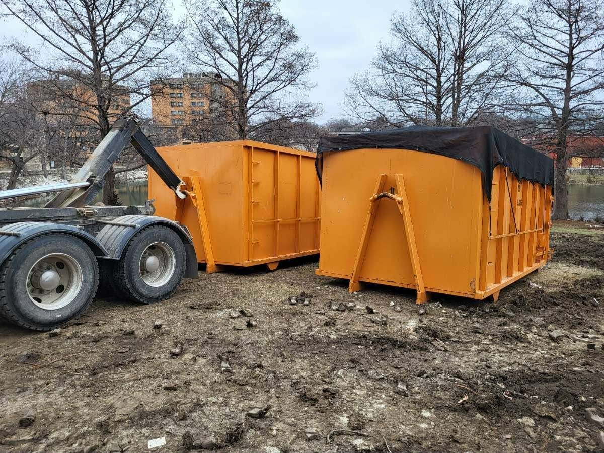 A roll-off truck is positioned to load a large, orange industrial dumpster in a dirt lot with bare trees in the background.