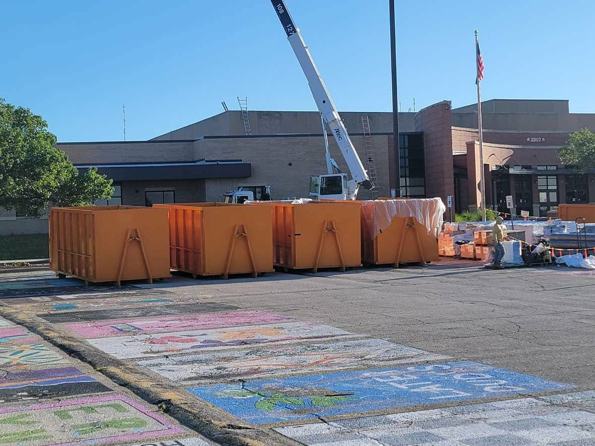 A crane stands before a building with a row of large, orange dumpsters in a parking lot featuring painted asphalt.