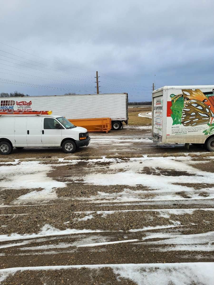 A white van and two parked trailers in a snowy, gravel lot under an overcast sky.