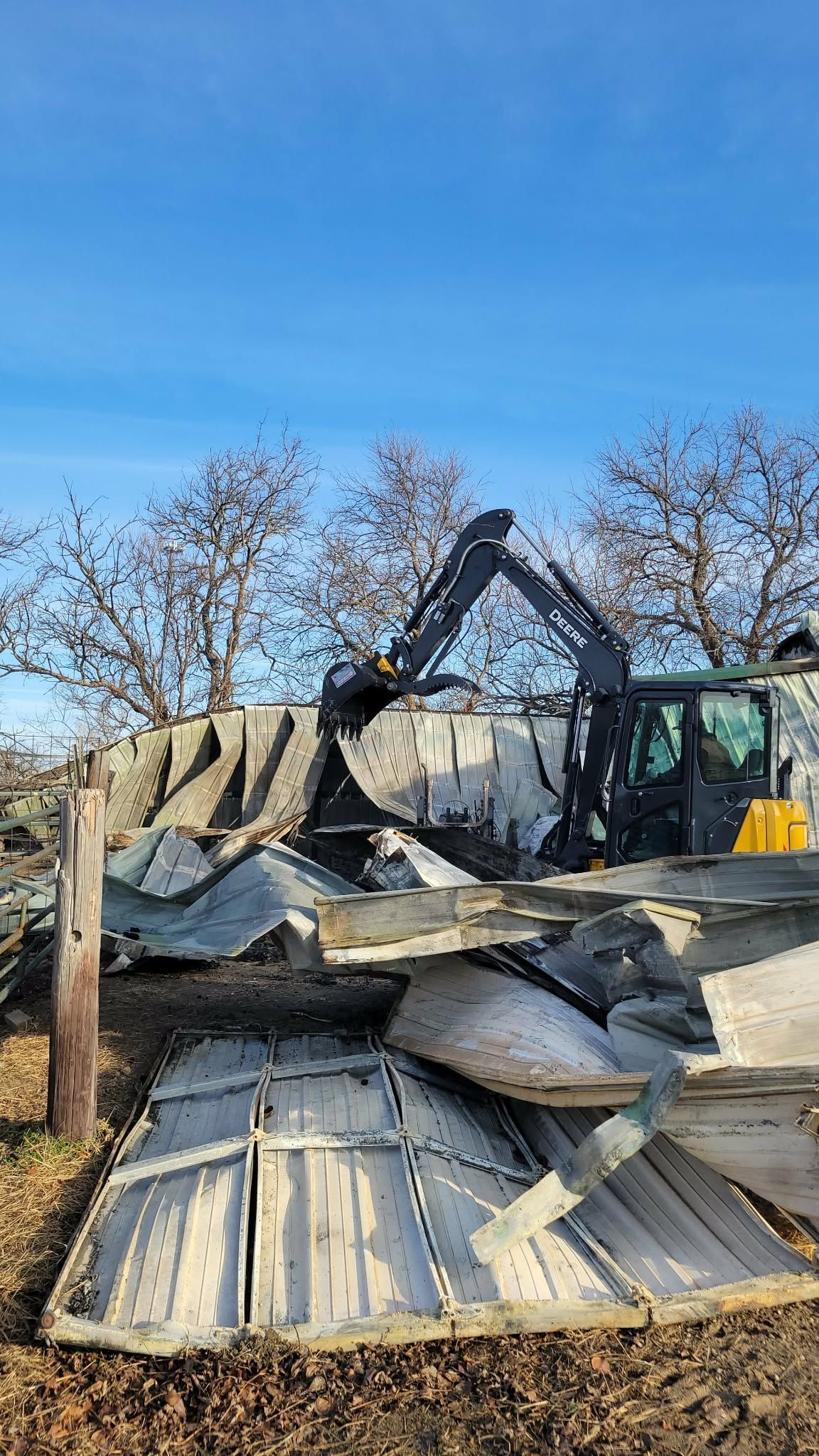 A dark-colored excavator demolishes a structure, moving sheet metal and debris in a field against a bright blue sky.