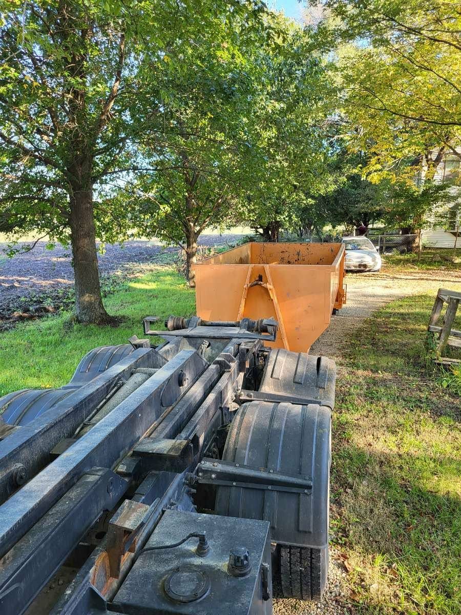 A view from behind a black roll-off truck chassis towards an orange dump container parked on a grassy path near trees.