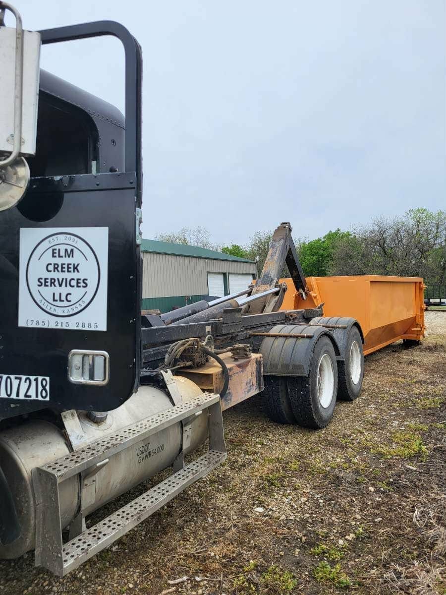 A black roll-off dumpster truck with an Elm Creek Services LLC logo, parked on a gravel lot near a building.