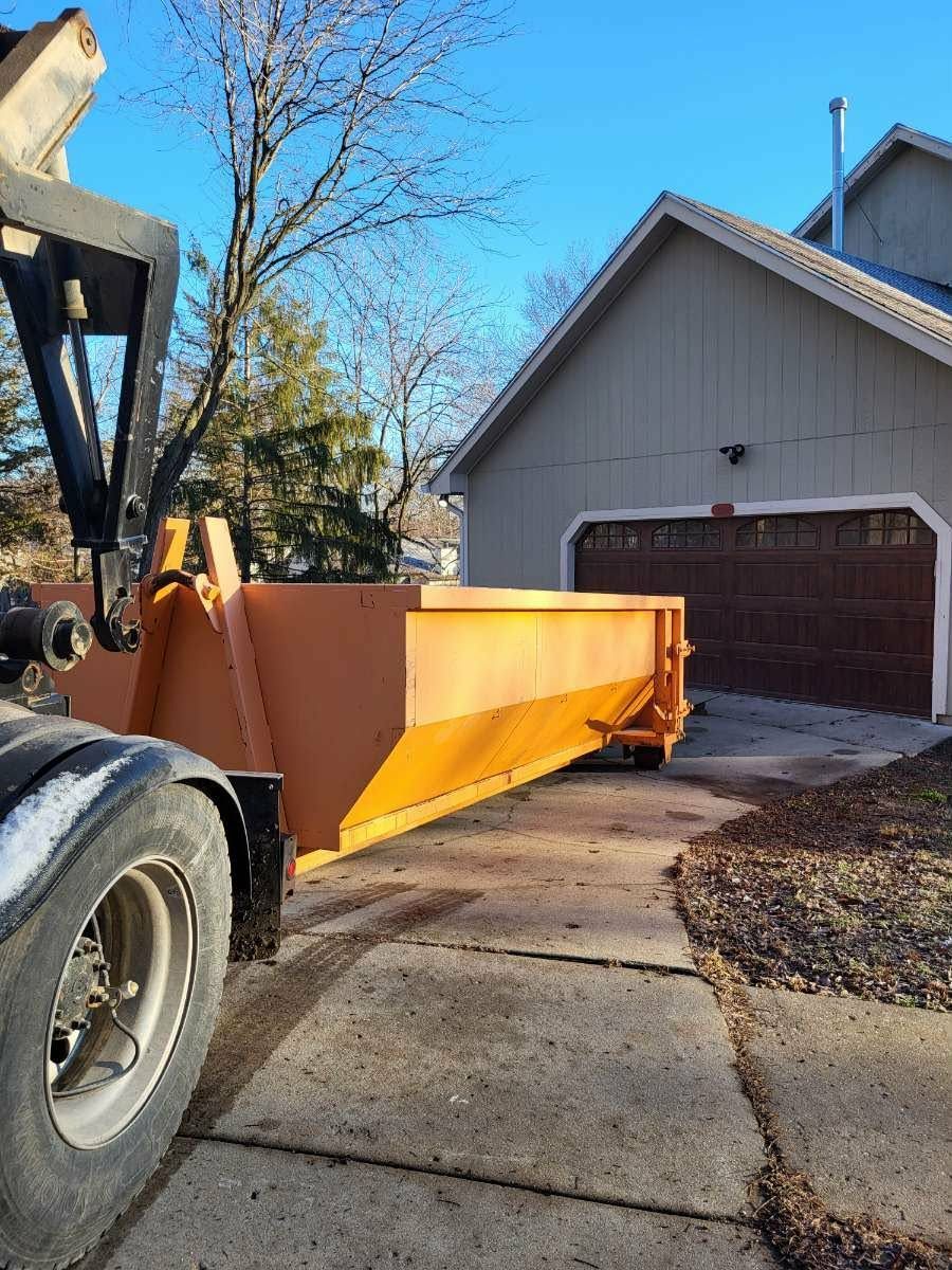 An orange dumpster being unloaded from a truck onto a concrete driveway in front of a residential garage.
