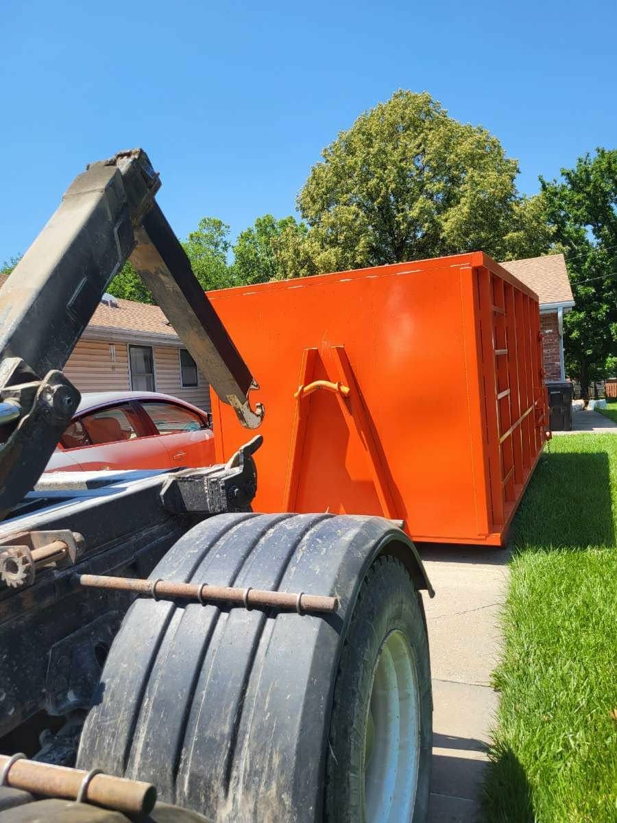 A bright orange roll-off dumpster sits on a driveway, attached to a delivery truck.