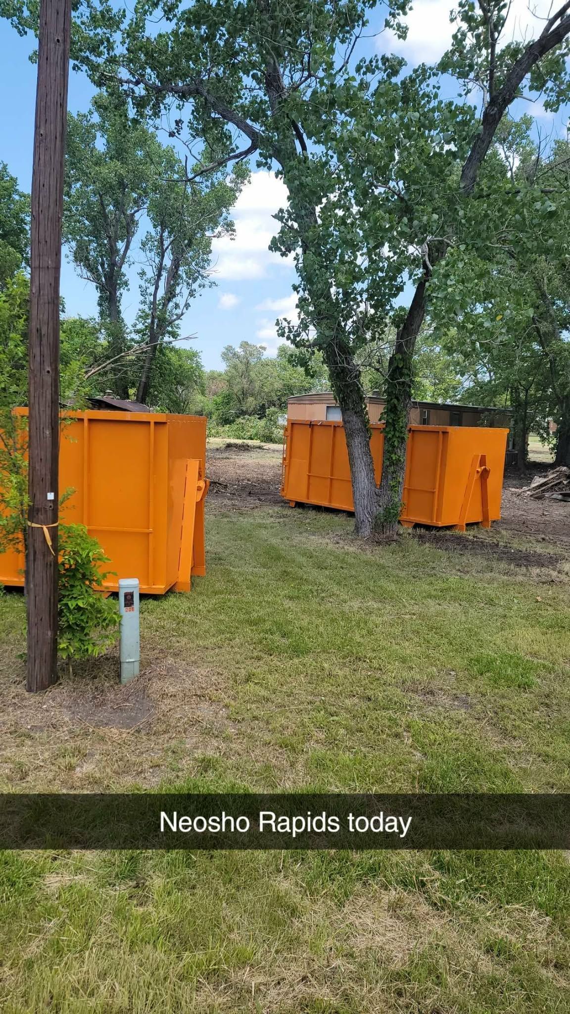 Two large, orange industrial dumpsters sit on a grassy area in a wooded clearing on a sunny day.