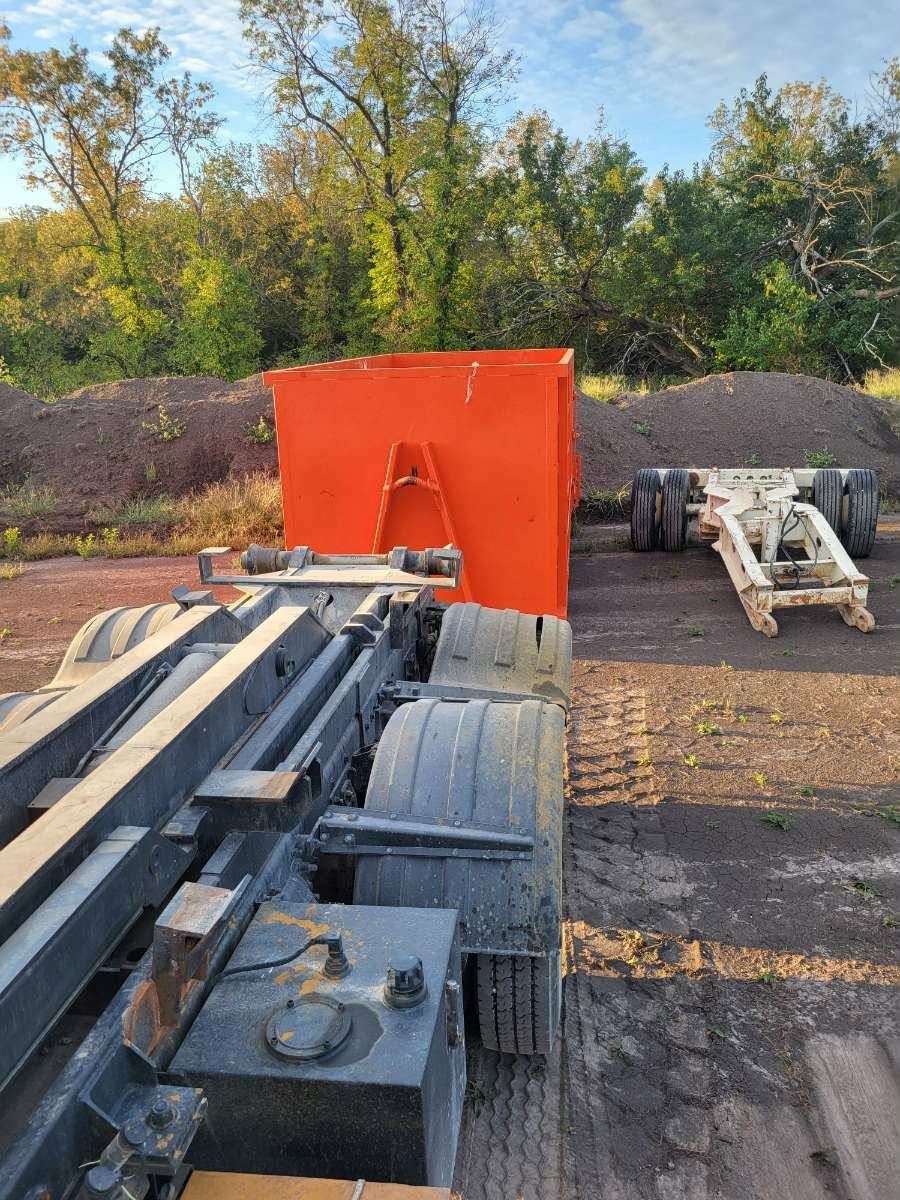A roll-off dump truck bed mechanism next to a bright orange container and a detached trailer frame in a gravel lot.