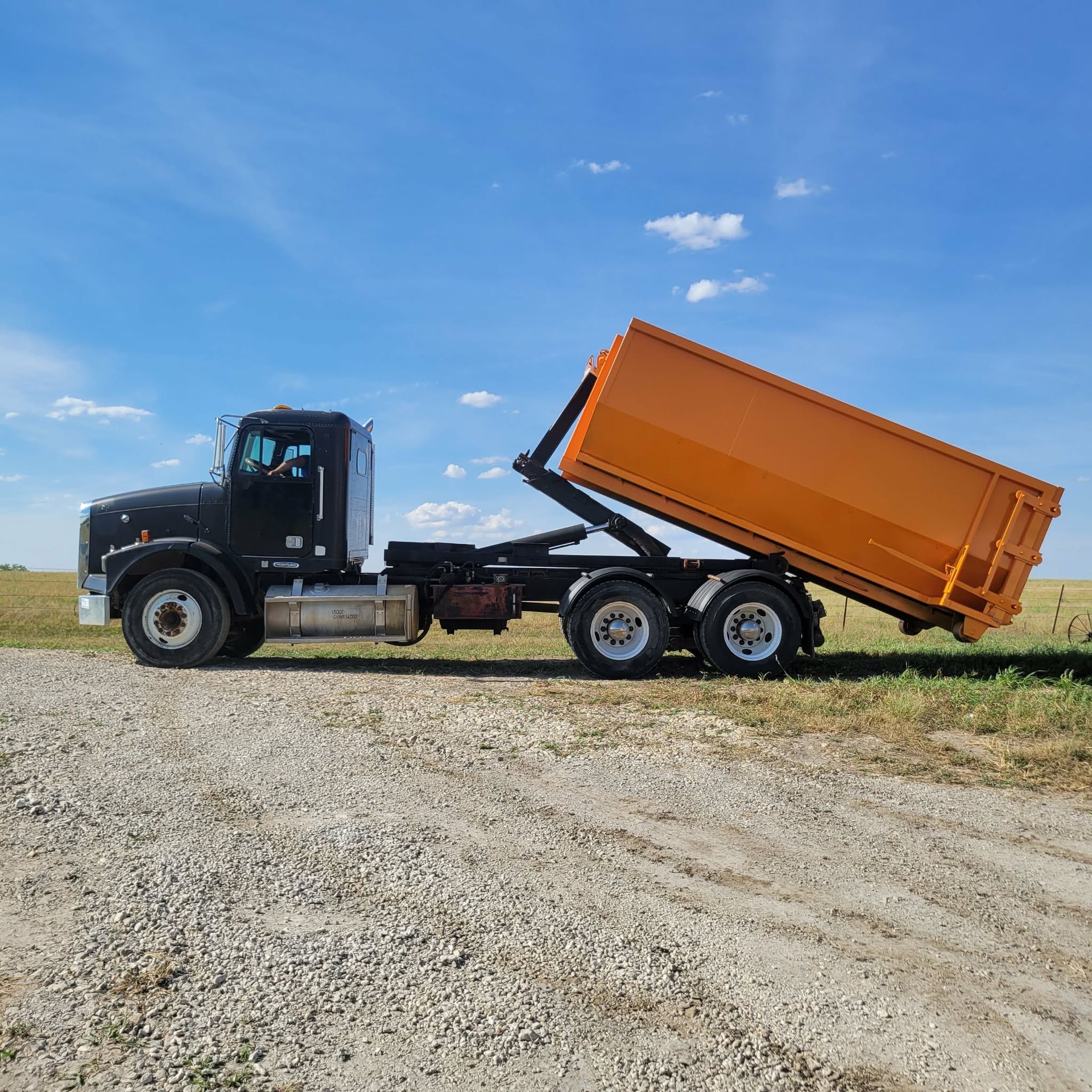 A black roll-off truck with a bright orange dumpster tilted upward, parked on a gravel road under a clear blue sky.