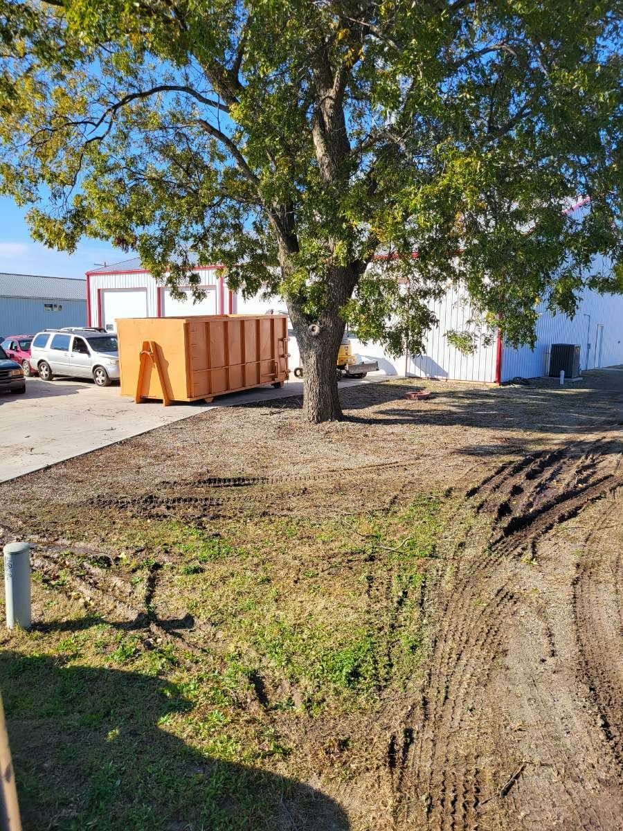 An orange dumpster sits under a large tree in a dirt lot next to a white industrial building and a silver van.