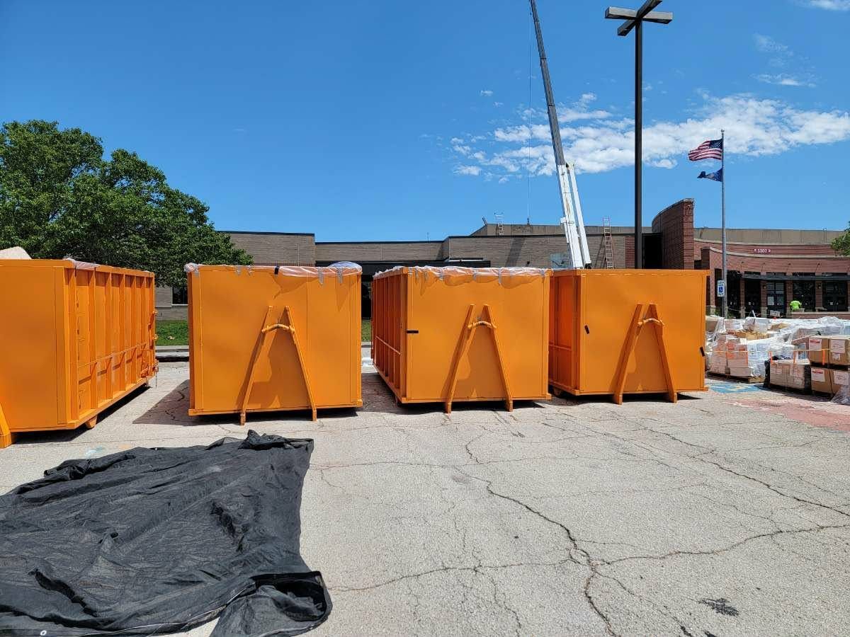 Four bright orange industrial dumpsters lined up in a sunny parking lot in front of a building with American flags.