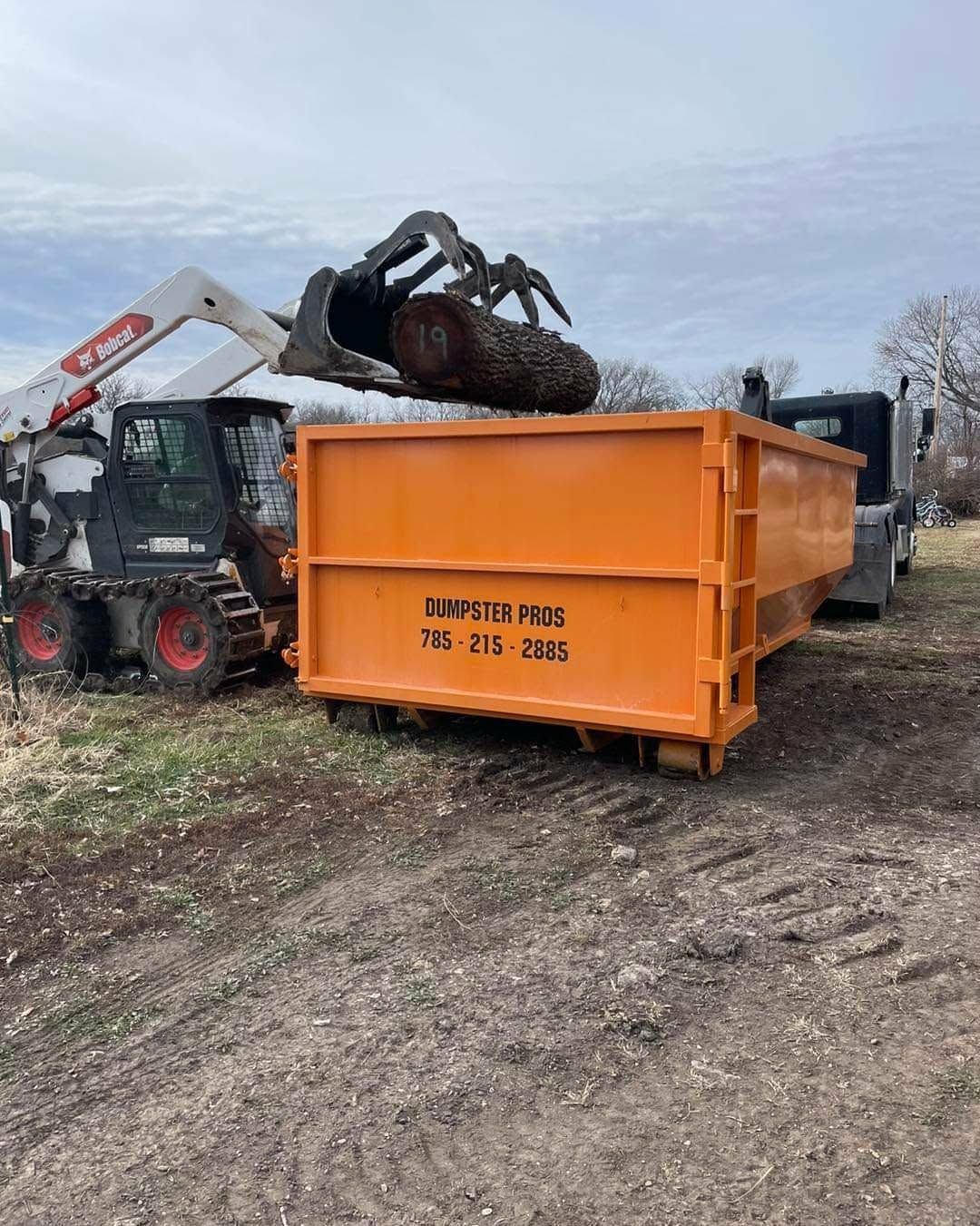 A skid steer with a grapple attachment loading a large log into an orange dumpster at a construction site.