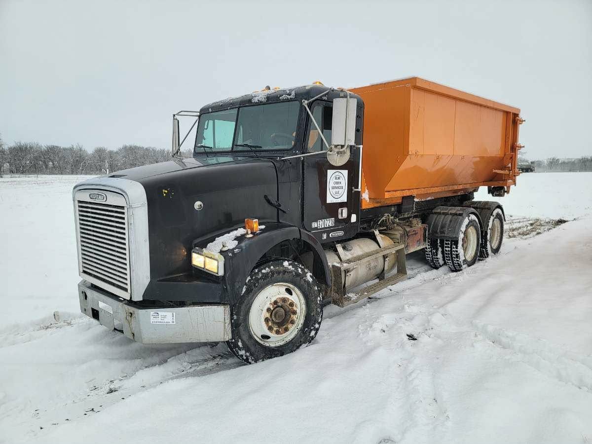 A black Freightliner dump truck with an orange container parked in a snow-covered field.