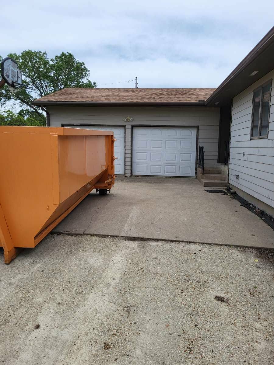 An orange dumpster sits in a driveway next to a residential garage and home exterior.