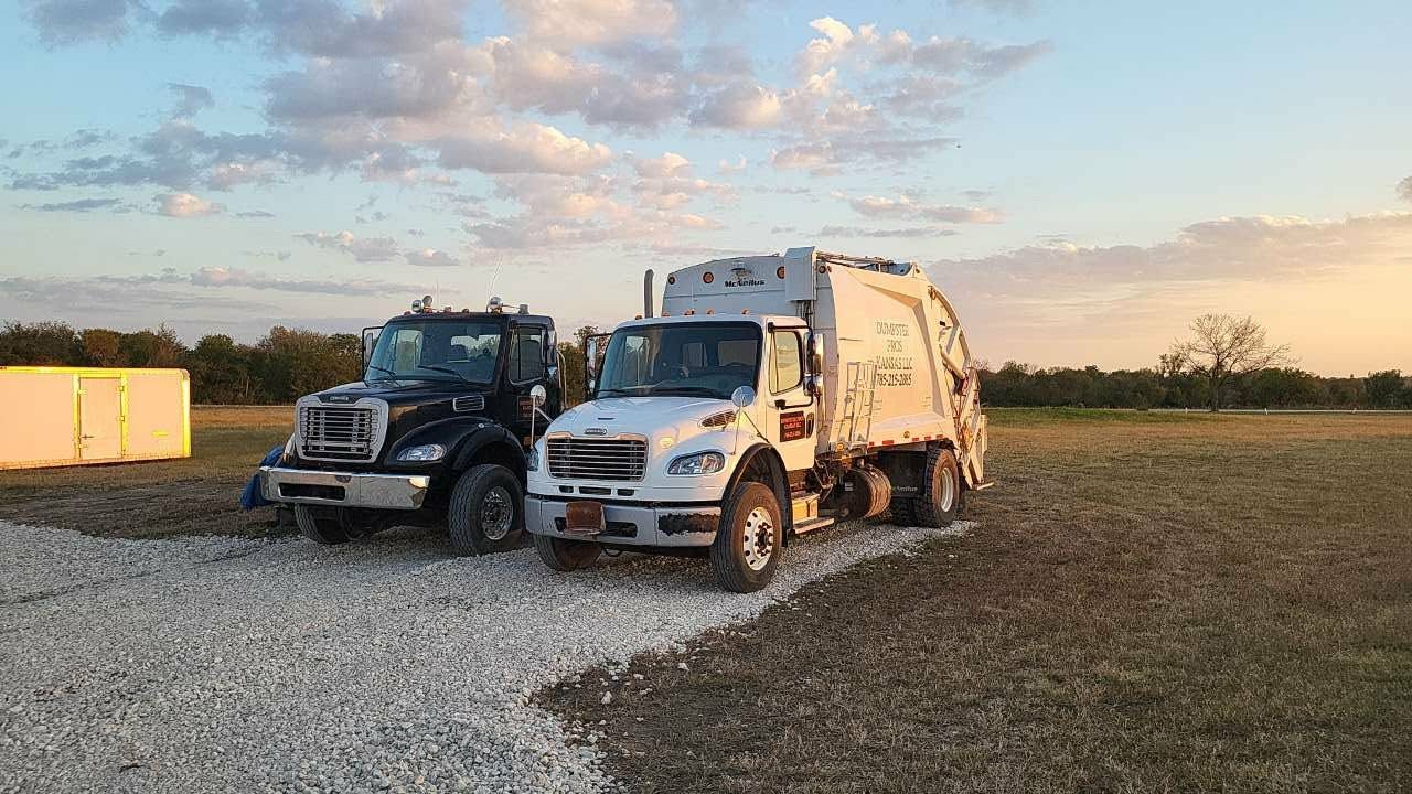 Two trucks, one black and one white garbage truck, parked on a gravel lot at sunset.