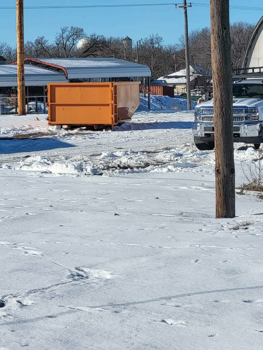 A snowy, sunlit scene featuring an orange dumpster in the background and a parked white truck near a wooden utility pole.