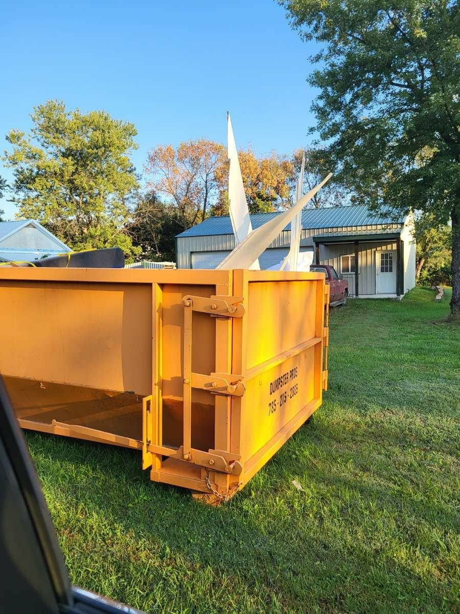 A large, empty, orange roll-off dumpster sits in a grassy yard near a garage and some trees under a clear blue sky.