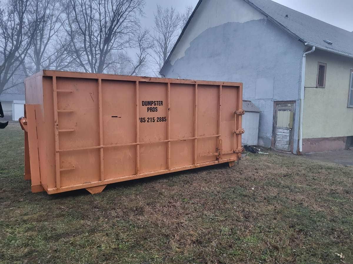 A large, empty orange metal dumpster sits on a grassy lot next to a light-colored building under an overcast sky.