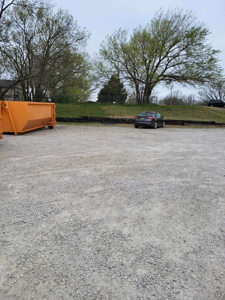 A gravel lot with an orange dumpster on the left and a dark sedan parked near a tree line in the background.