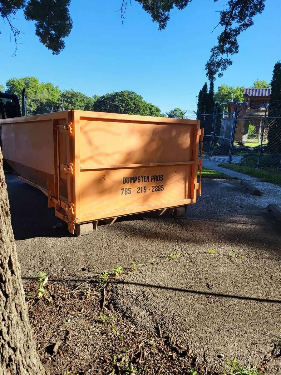 An orange roll-off dumpster parked on a gravel surface outdoors with a phone number and company name printed on the side.