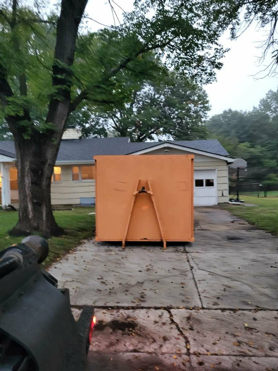 An orange dumpster sits in a residential driveway in front of a house with a garage and a large tree.