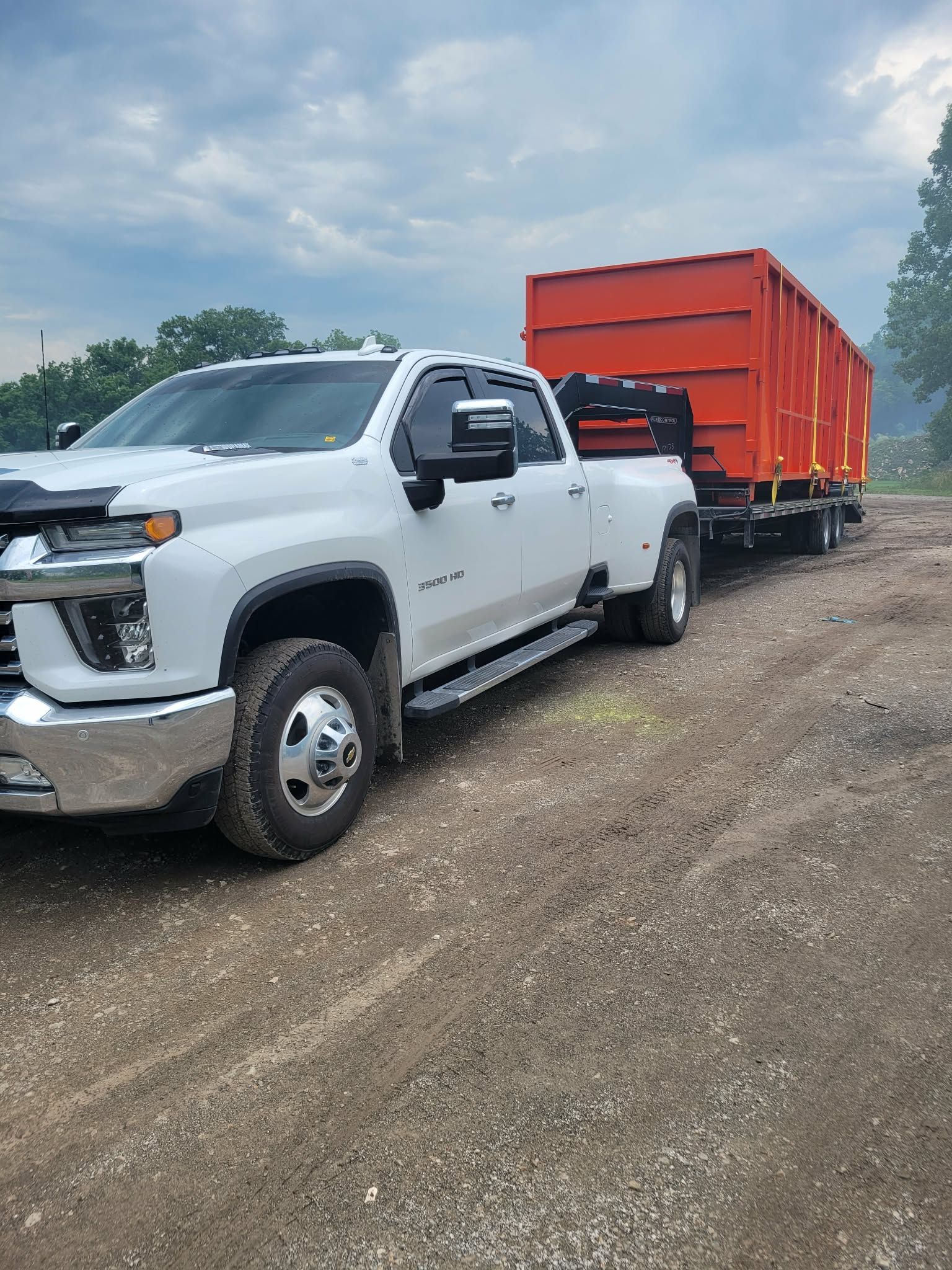 A white Chevrolet dually pickup truck towing a large orange industrial dumpster on a trailer across a gravel lot.