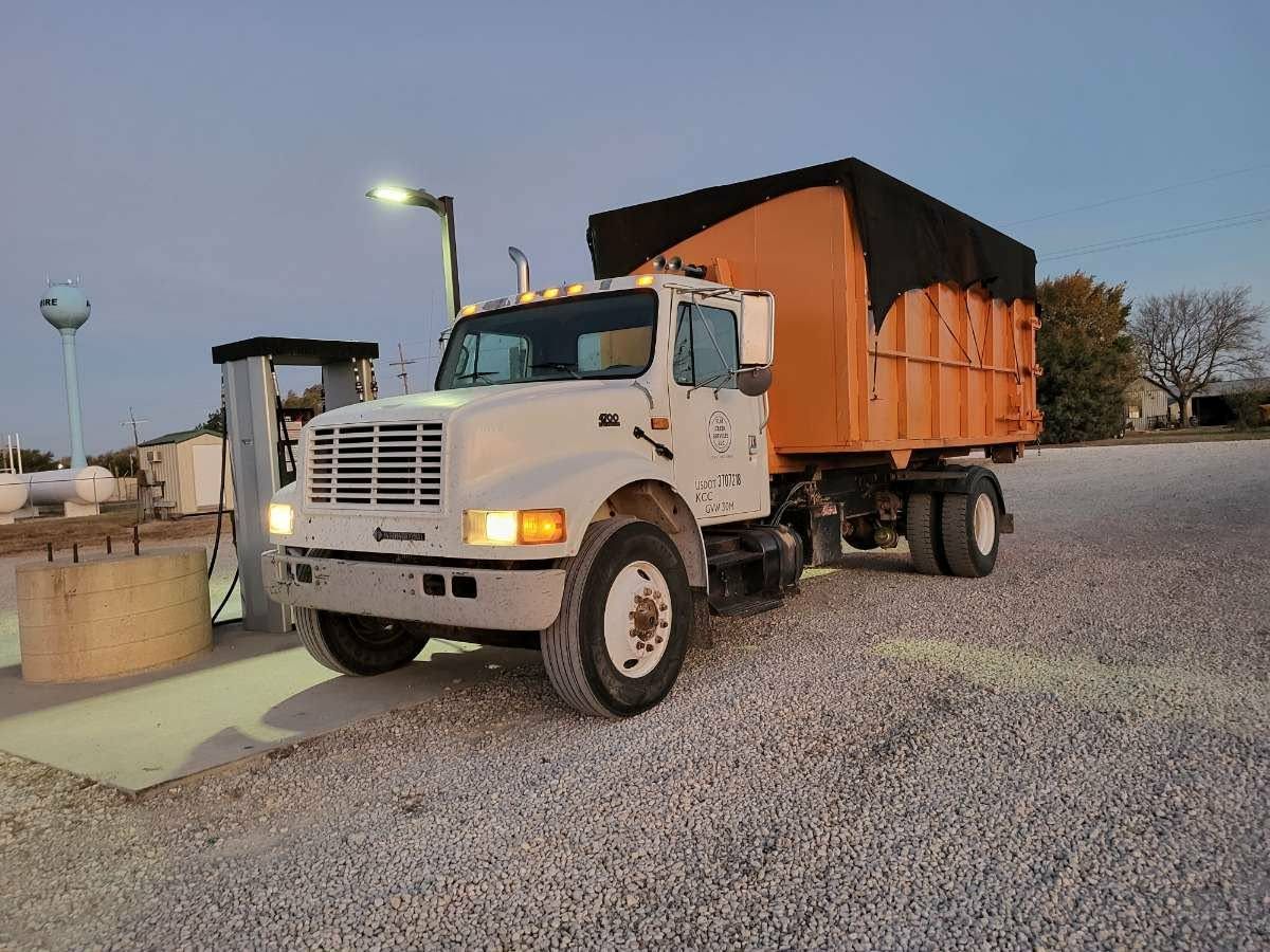 A white commercial dump truck with an orange container parked on a gravel lot next to a fuel pump at twilight.