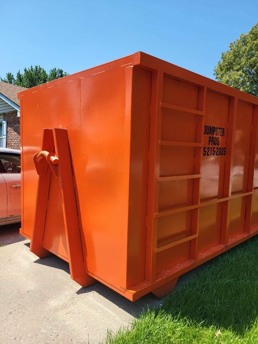 A bright orange metal roll-off dumpster sits on a residential driveway next to a grassy lawn under a clear blue sky.