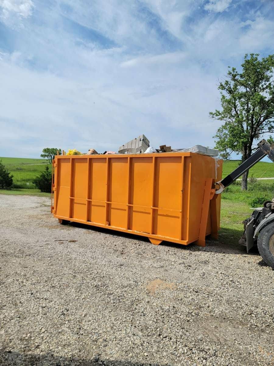 A bright orange roll-off dumpster sits on a gravel lot under a blue sky, partially attached to a truck.