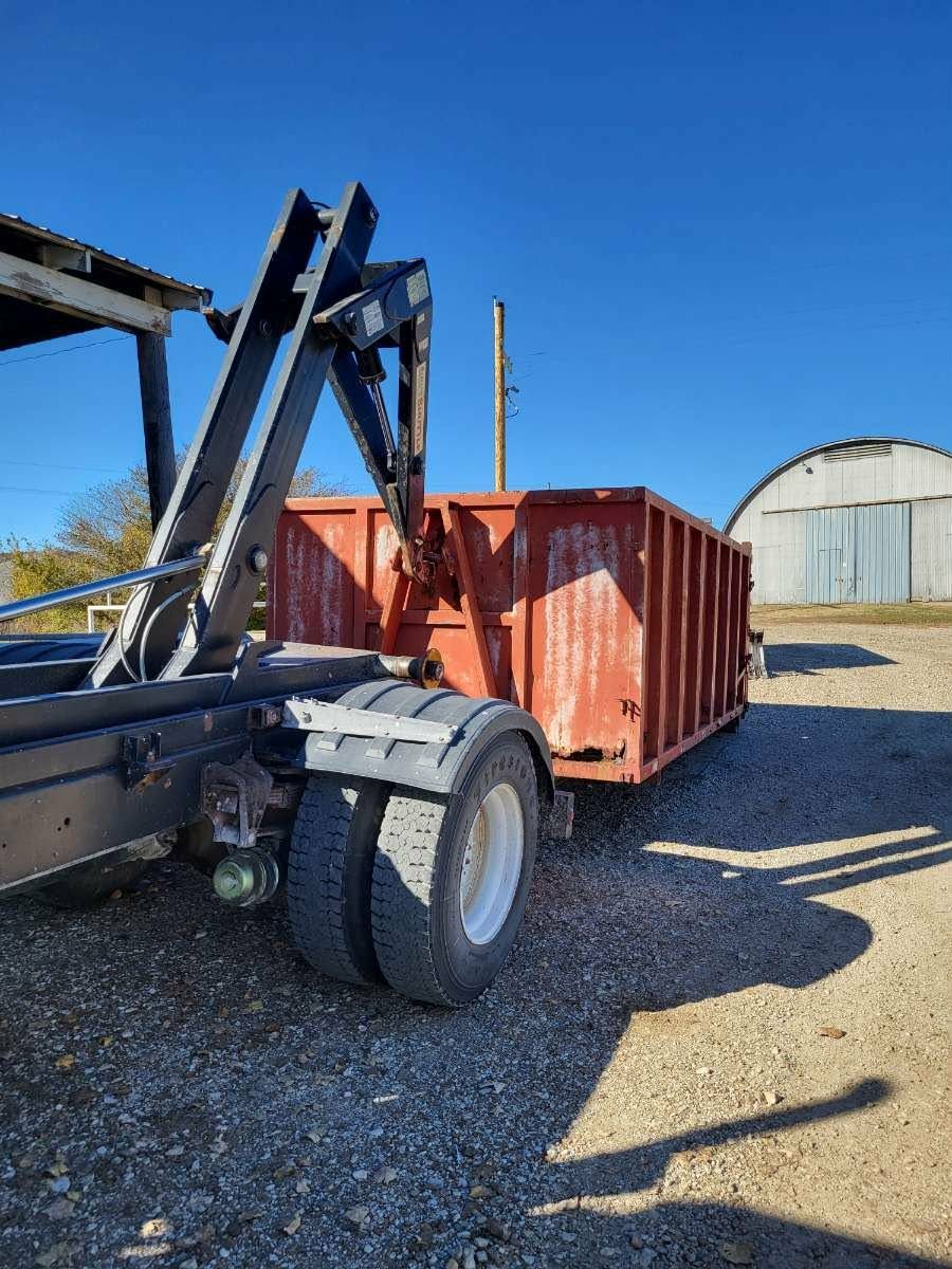 A hooklift truck loading a large, rusted red metal container onto its frame outdoors on a sunny day.