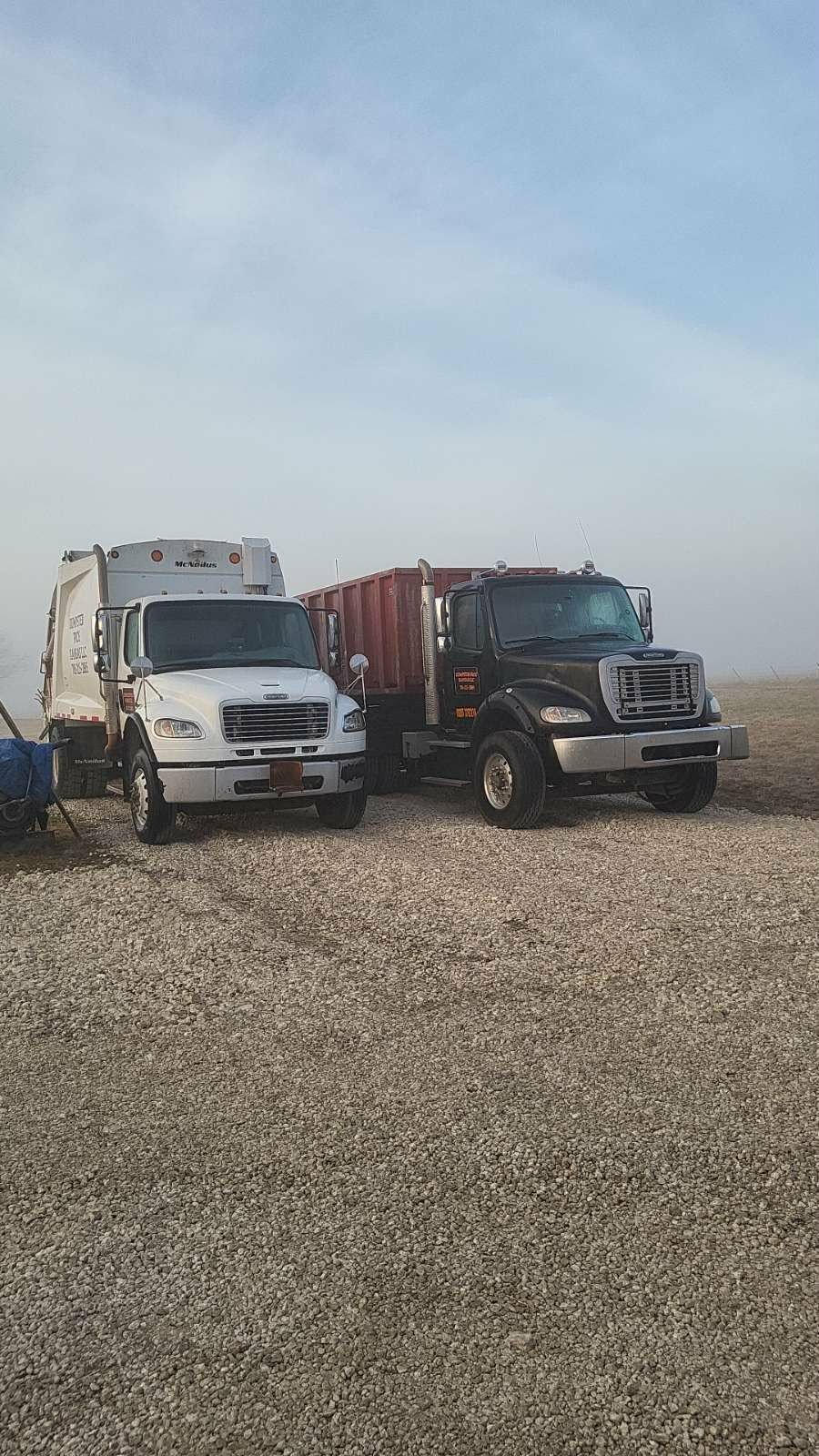 A white garbage truck and a black roll-off dumpster truck parked on a gravel lot under a pale, hazy sky.