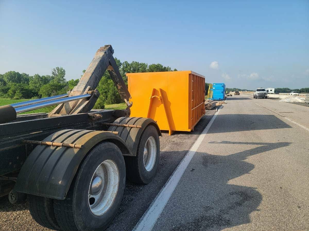 A large yellow industrial dumpster being loaded onto the back of a truck on the side of a paved road.