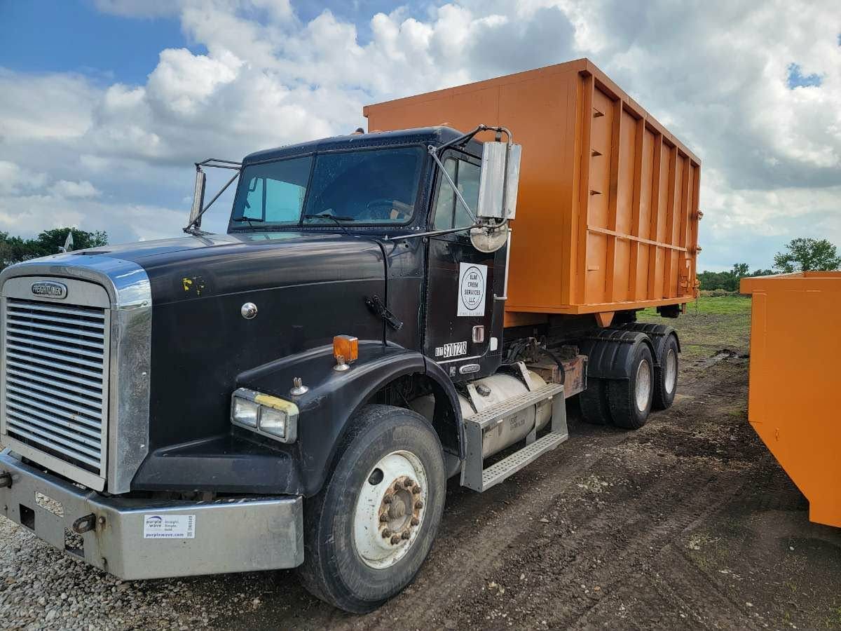 Black Freightliner roll-off truck with a large orange waste container parked on a dirt lot under a cloudy sky.