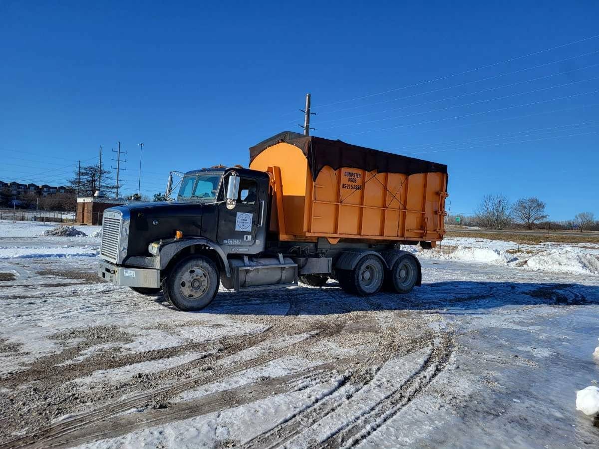 A black commercial truck with a large, orange, covered roll-off dumpster parked on a snow-covered, icy lot under blue sky.