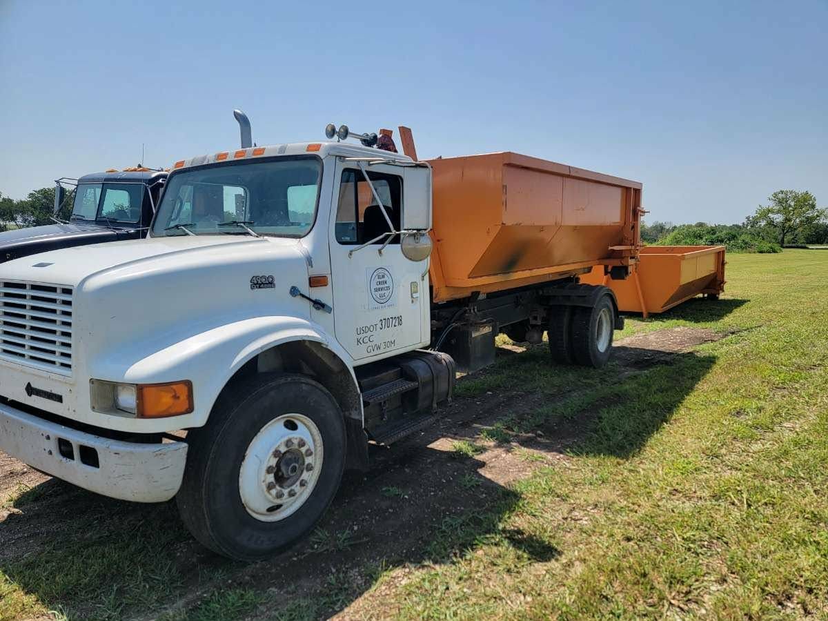 A white dump truck with an orange trailer parked on a grassy field under a clear blue sky.