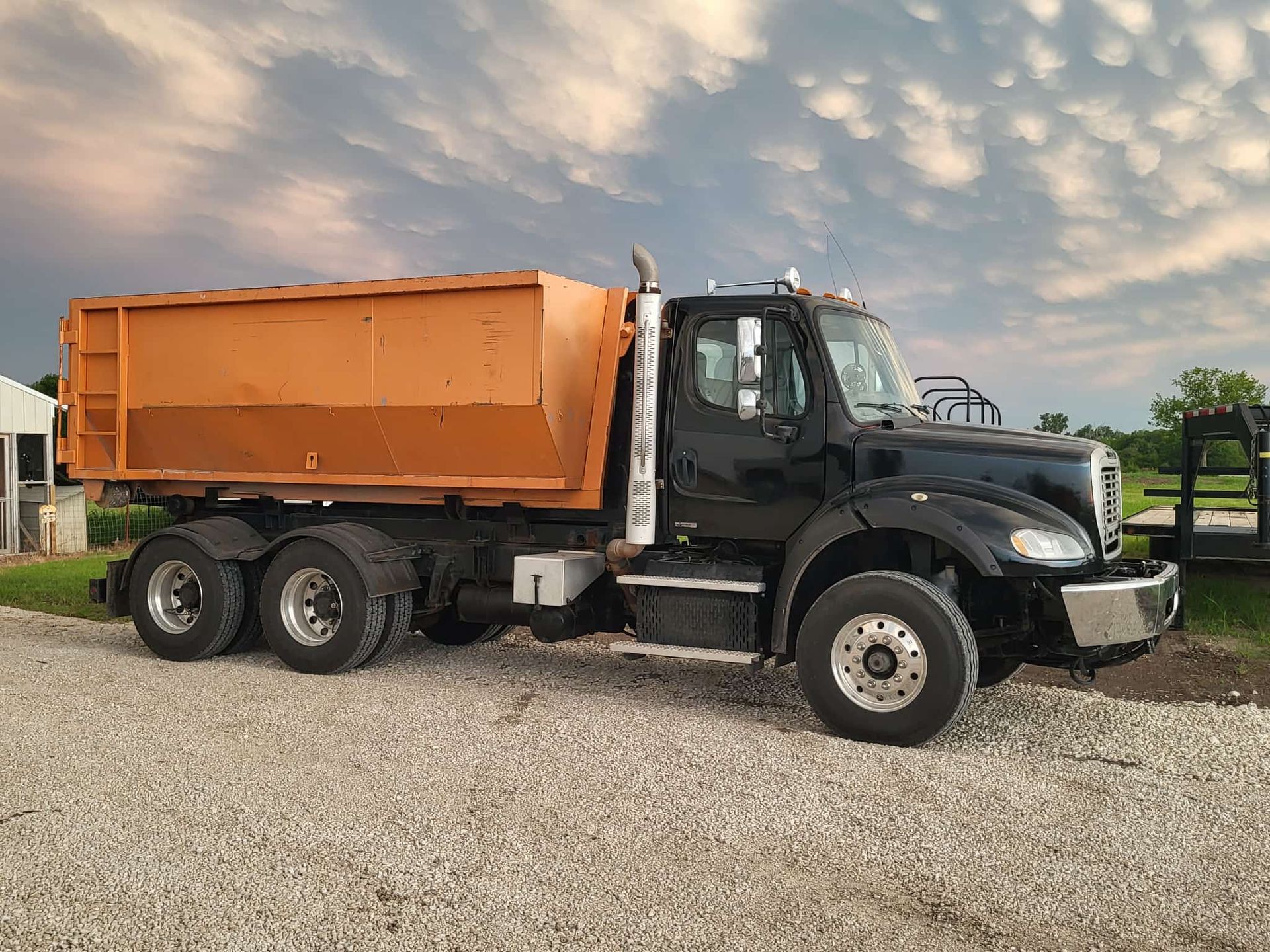 A black dump truck with an orange container parked on a gravel lot under a cloudy sky.