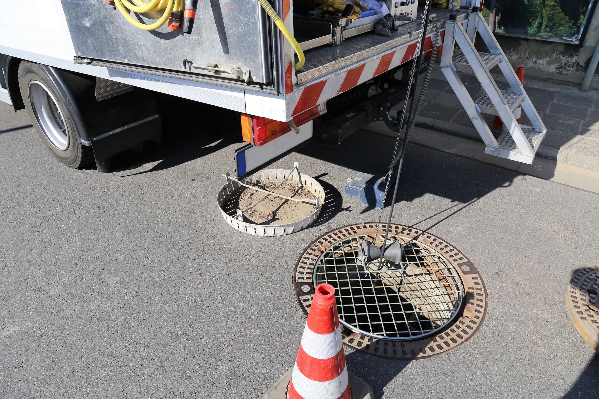 Camion da cantiere stradale sopra un tombino aperto con cono di sicurezza e scala metallica sul marciapiede