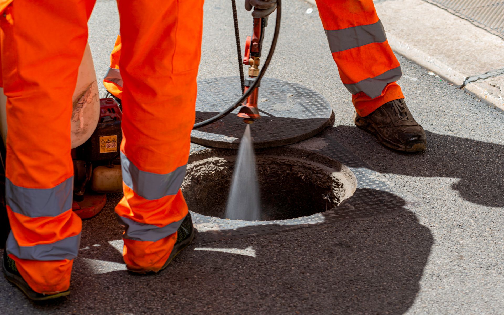 Gli operai, vestiti con indumenti di sicurezza arancioni, tagliano l'asfalto intorno a un tombino con un getto d'acqua.