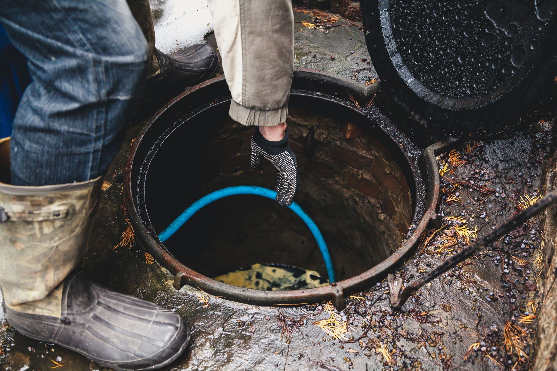 Un operaio usa un tubo flessibile per pulire un tombino o un'apertura fognaria con acqua fangosa e stivali nelle vicinanze.
