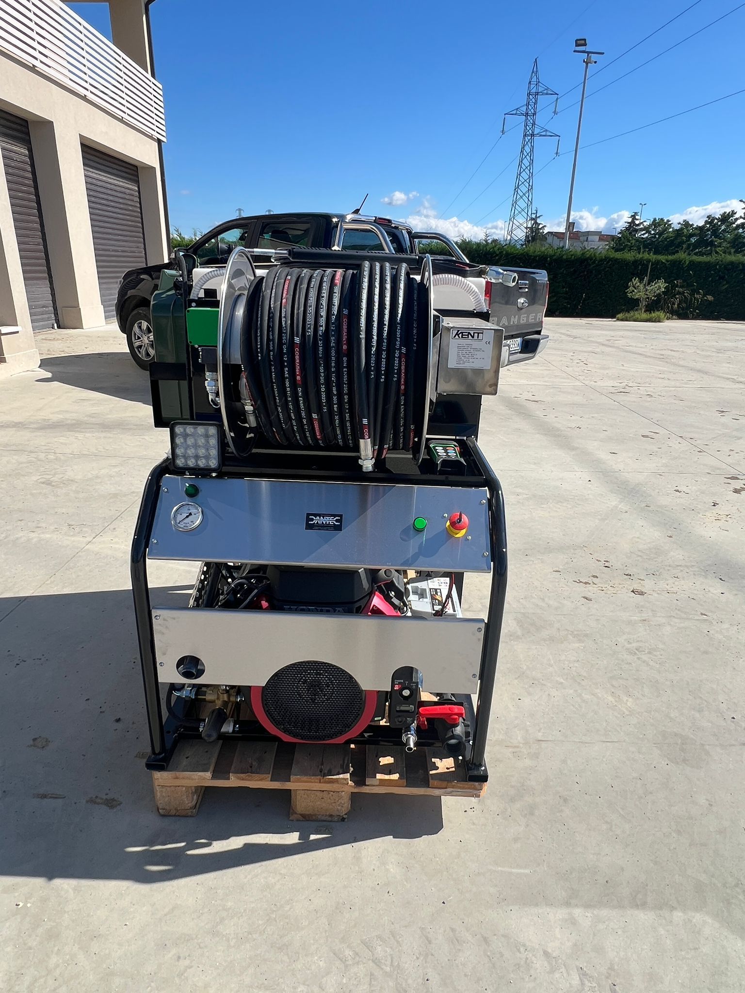 Compressore industriale su pallet all'esterno di un edificio, con motore nero su un serbatoio grigio e tubi flessibili collegati.