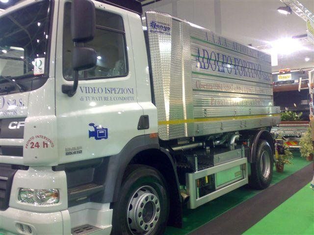 Camioncino bianco per le consegne esposto al chiuso in una mostra, con finiture e scritte verdi sul lato.