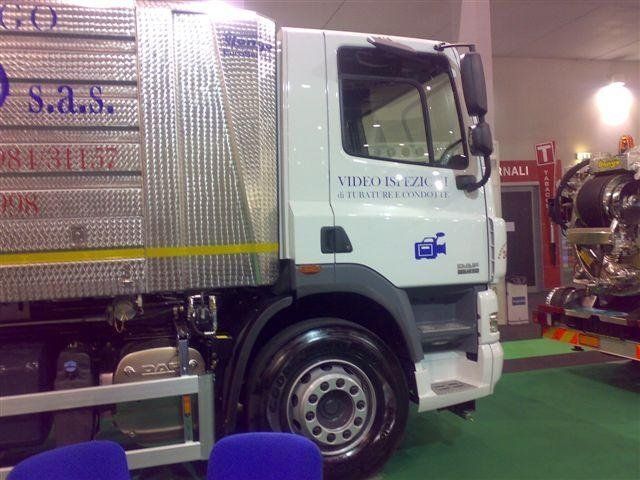 Cabina di camion bianca con cassone argentato esposta al coperto su un tappeto verde in occasione di un salone dell'auto.