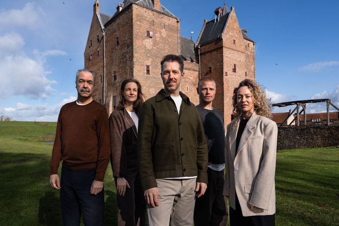 Vier mensen staan ​​op het gras voor een groot stenen kasteel onder een blauwe hemel.