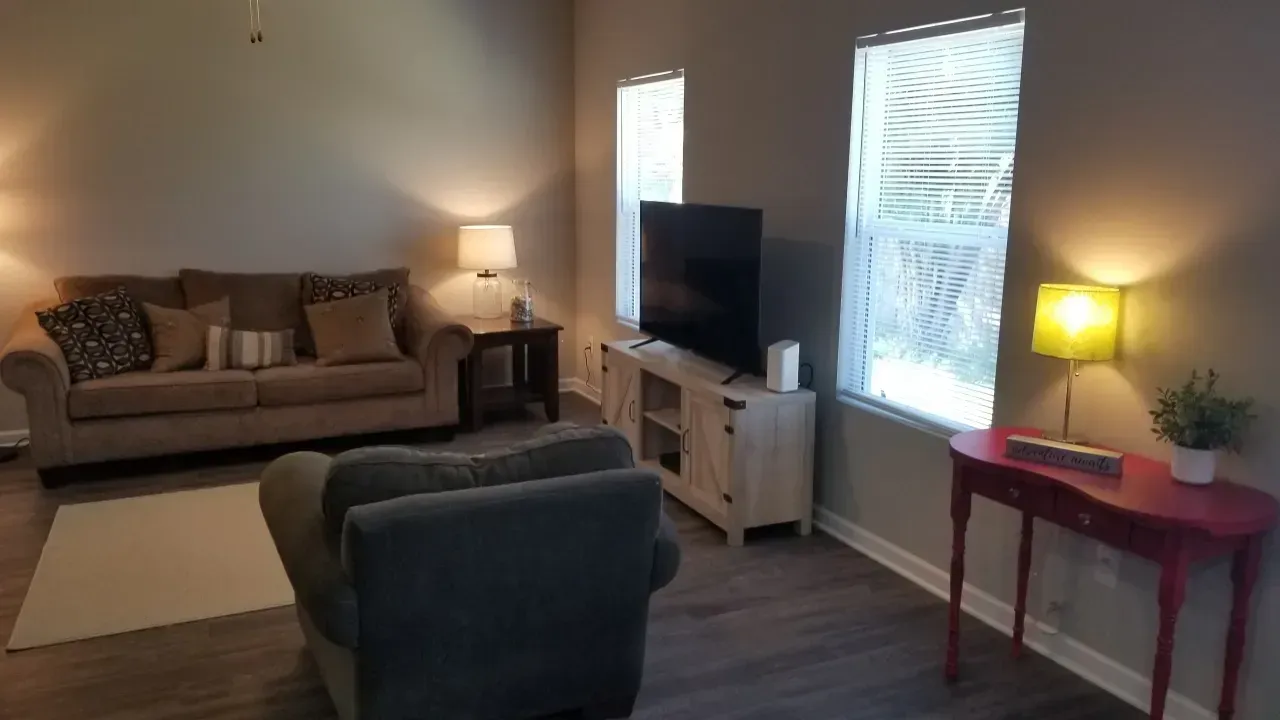 Cozy living room with sofa, armchair, TV, two lamps, and a red side table by windows