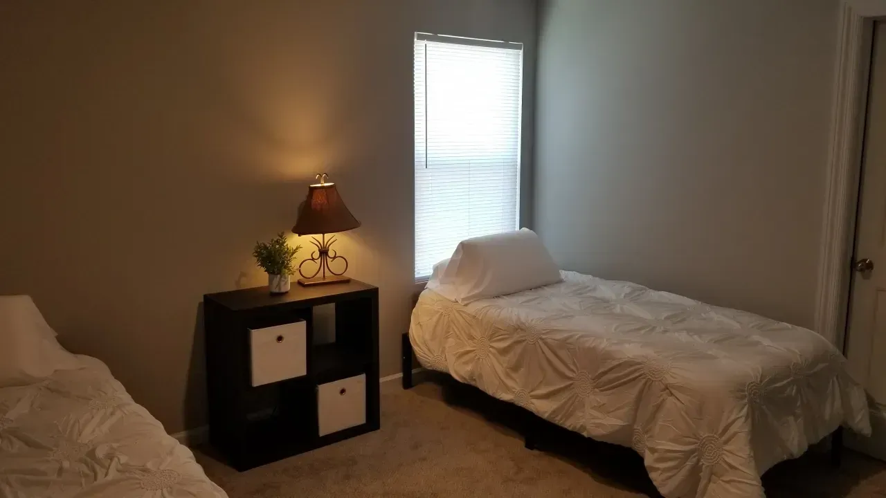 Simple bedroom with two single beds, a nightstand lamp, and a window with blinds.
