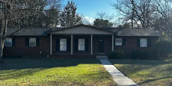 Single-story brick house with a front porch and walkway, set on a sunny lawn.
