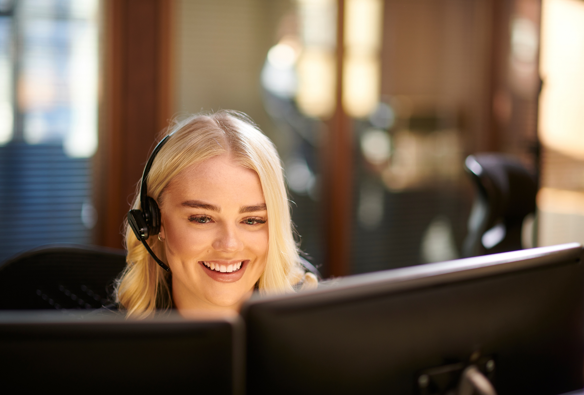 Blonde woman wearing a headset smiles at a computer screen in an office.