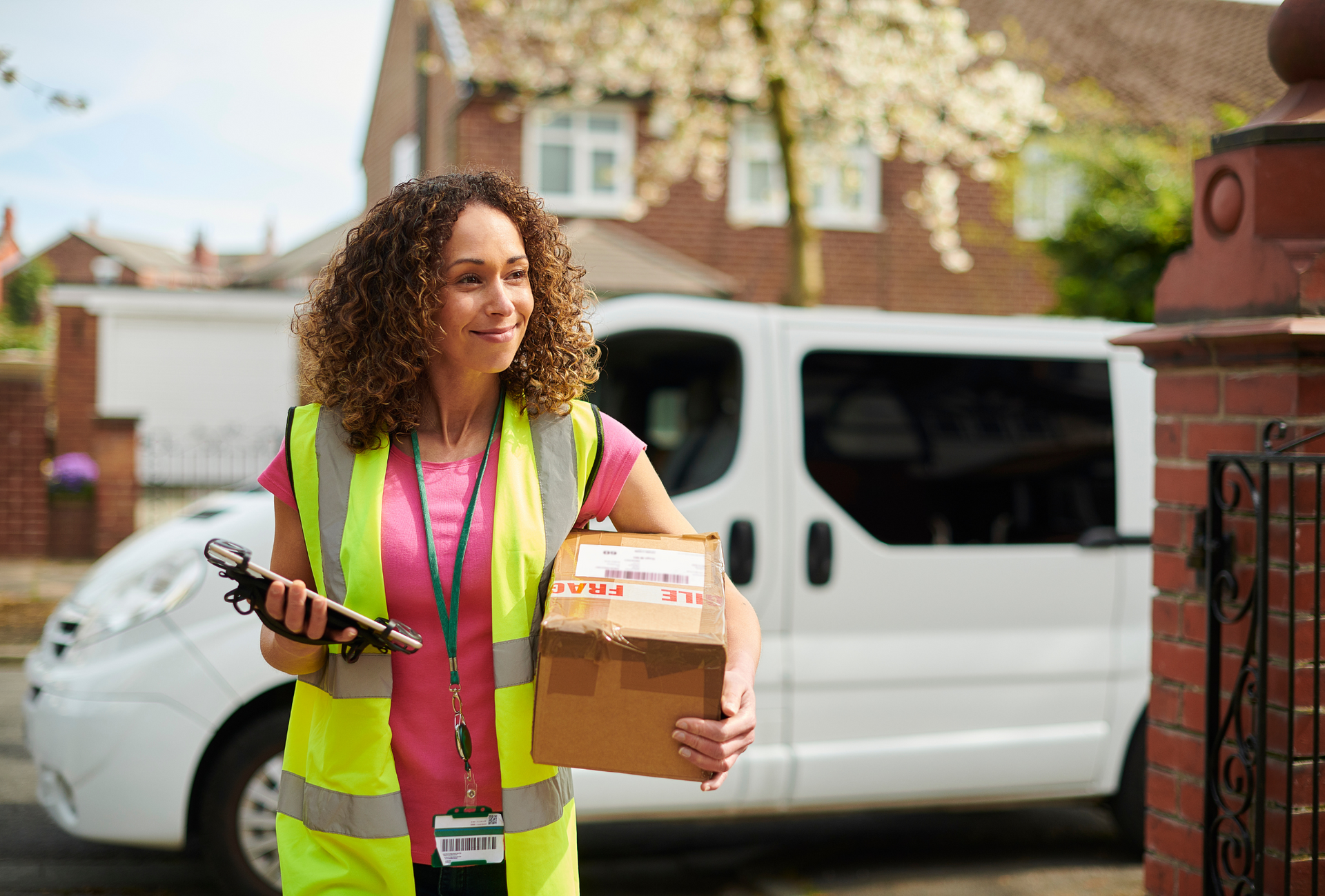 Delivery person with curly hair smiles, holding a package and scanner, beside a white van in front of a house.