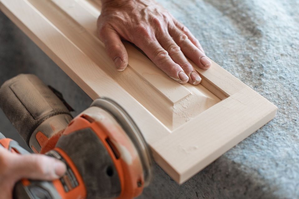Hands sanding a white wooden frame with an orbital sander on a workbench