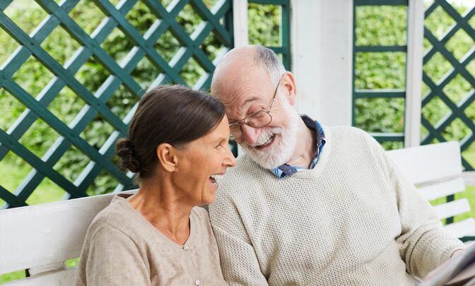 Elderly couple laughing together on a white bench, green background.