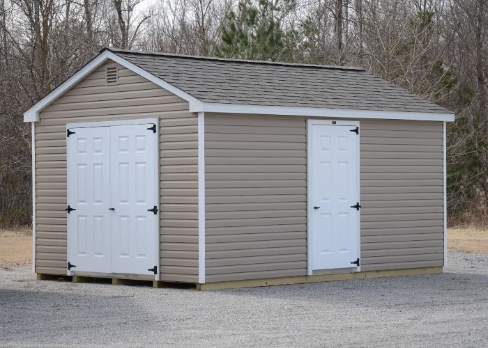 Tan storage shed with white double doors and a side entry door on a gravel lot near trees